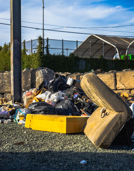 A waste collection worker wearing a high-visibility yellow and red uniform is operating a large red rubbish truck on the side of a street. The rear loading mechanism of the truck is open, revealing in