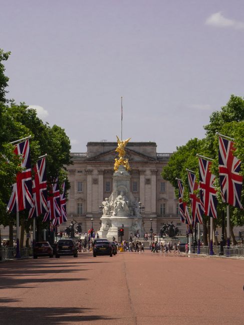 A wide street lined with tall trees on both sides, decorated with numerous Union Jack flags attached to poles along the pavement. The flags flutter gently in the breeze, creating a colorful and patrio