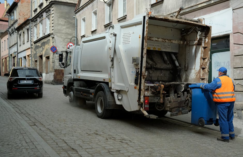 A worker dressed in a yellow high-visibility vest and black pants is operating a red rubbish collection truck on the side of a road, with the rear compartment open and filled with waste. The worker ap