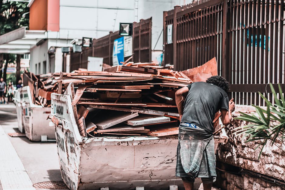 A man with curly dark hair, wearing a dark T-shirt and a plaid kilt, is standing with his back to the camera next to a large, overflowing skip filled with discarded wooden planks, tiles, and construct
